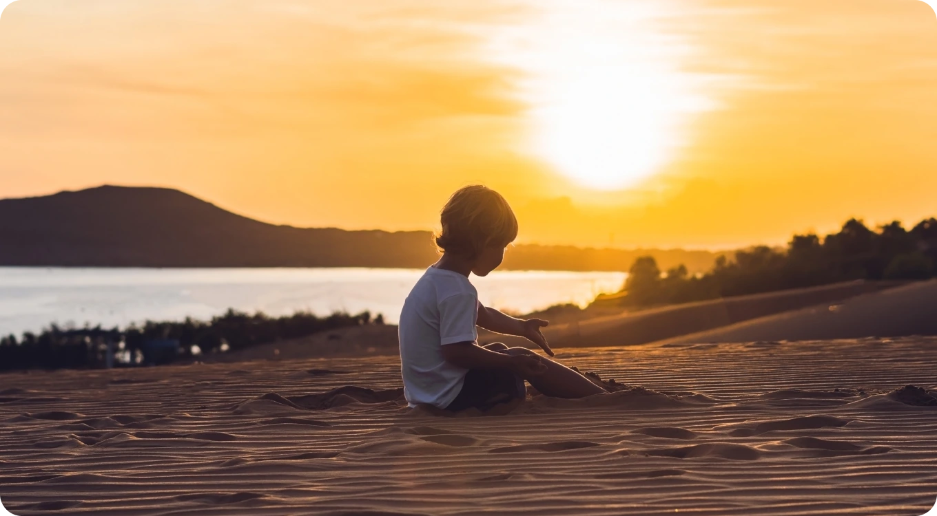 A young child sits on rippled sand dunes at sunset, looking toward a calm body of water and distant hills. The golden sky and low sun create a peaceful silhouette scene with warm evening light.