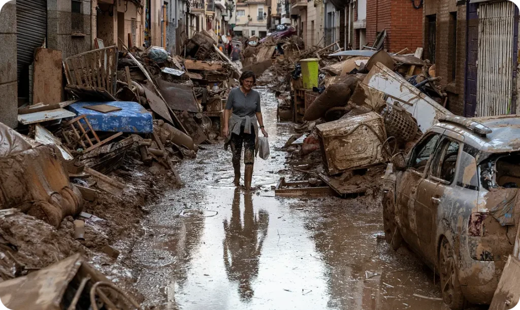 A person walking through a flooded street filled with mud, debris and storm-damaged cars.