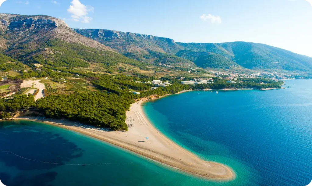 Aerial view of a curved sandy beach reaching into the turquoise Adriatic Sea, backed by green hills.